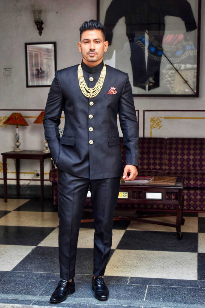 Man in traditional black Jodhpuri suit with necklace posing in an elegant living room.
